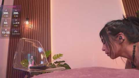 A video of a woman with dark hair, glasses, and a tattoo, looking at a digital thermometer on a wooden wall.