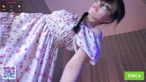 Video of a young woman with pale skin, black hair, and glasses, wearing a white floral dress, in a dimly lit room.