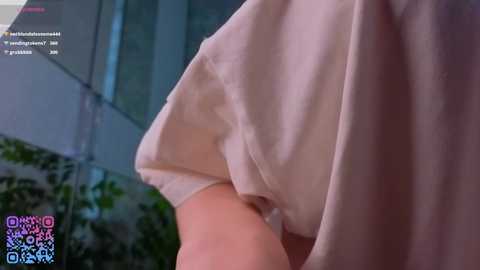 Video of a light-skinned person wearing a white t-shirt, leaning against a modern, glass-walled building. The person's arm and part of the torso are visible. A QR code is partially visible in the bottom left corner.