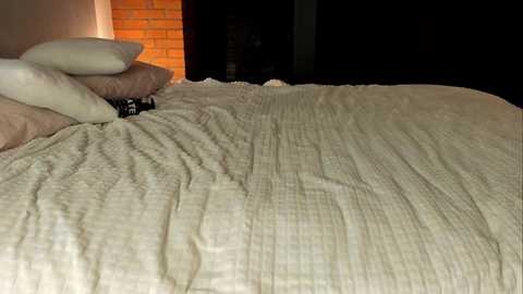 Video of a neatly made bed with a textured, cream-colored quilt and two white pillows. A brick wall and dark, unlit room are in the background, creating a cozy, warm ambiance.