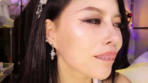 Close-up video of a woman with fair skin, dark hair adorned with silver headpiece, wearing intricate silver earrings. Background shows indoor setting with blurred objects and soft lighting.