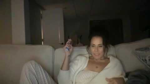 Video of a dark-haired woman in a white robe, holding a cigarette, sitting on a beige couch in a dimly lit living room.