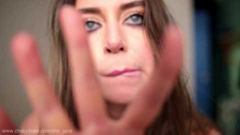 Video of a young woman with fair skin, blue eyes, and long brown hair, wearing pink lipstick, making a \"come here\" gesture with her right hand. Background is blurred.