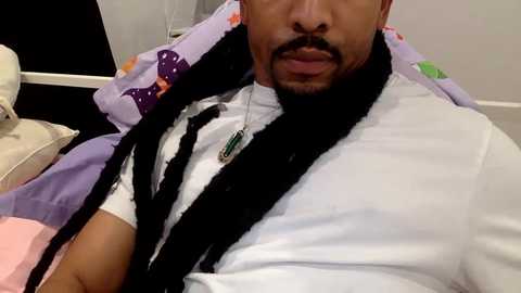 Video of a man with a mustache and beard, wearing a white shirt, black necklace, and black braids, reclining on a purple bed with a white pillow.