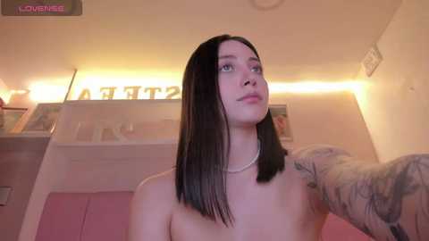 Video of a topless young woman with straight, dark hair, blue eyes, and light skin, sitting in a dimly lit room with pink walls, a white ceiling, and a framed \"LOVE\" sign.