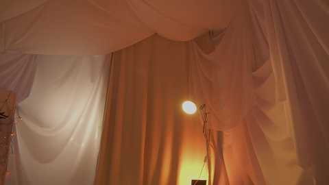 Video of a cozy indoor scene with soft, warm lighting. White sheer curtains draped over a canopy bed, creating a romantic ambiance. A small lamp casts a gentle glow, adding a cozy, intimate feel.