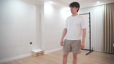 A video of a young, fair-skinned man with dark hair, wearing a white t-shirt and beige shorts, standing in a minimalist, white-walled room with a wooden floor, a black exercise rack, and light beige curtains.