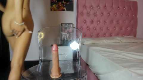 Video of a nude woman with a medium-sized, erect penis, standing beside a clear acrylic chair with a flesh-colored dildo on a glass table in a pink-tufted bedroom.