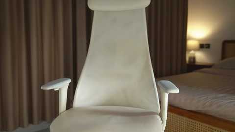 Video of a modern, white ergonomic chair with a high backrest and armrests, positioned in a dimly-lit bedroom with a bed, wooden nightstand, and beige curtains.