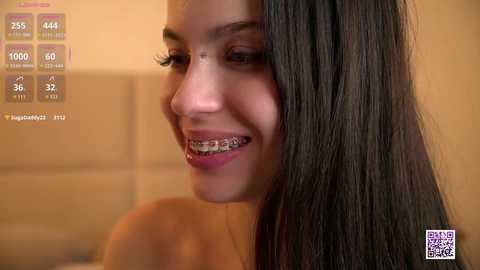 Video of a smiling, light-skinned woman with straight, long brown hair, wearing braces, in a bathroom with beige tiles.