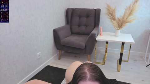 A video of a light-skinned woman lying on a black mat, wearing a beige top, with a gray armchair and white table with gold legs, featuring a vase of dried pampas grass in a minimalist room.