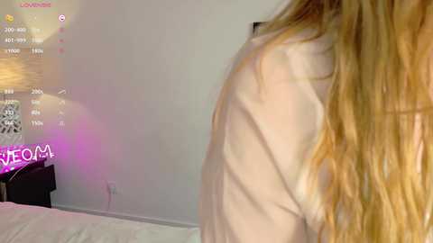 Video of a woman with long, blonde hair, wearing a white top, standing in a minimalist, modern bedroom with white walls, a pink light, and a TV displaying \"LIVE\" and \"ROOM.\