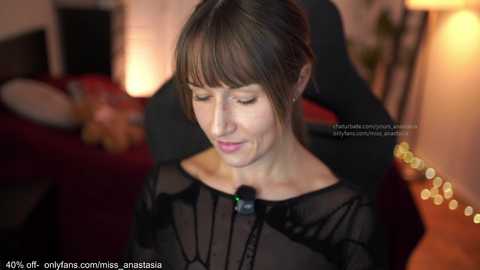 Video of a woman with dark hair, wearing a black sheer top, sitting on a black chair, in a dimly lit room with warm lighting and blurred background objects.