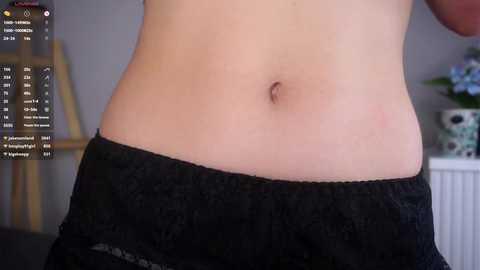 Video of a fair-skinned person's bare midriff in black shorts, with a blurred background featuring a white radiator and floral decor.