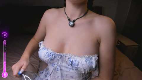 Video of a fair-skinned woman with a medium build, wearing a strapless, lacy, blue and white dress, a black necklace with a poker chip pendant, holding a thermometer, indoors on a bed with dim lighting.