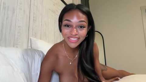 Video of a topless Black woman with long black hair, smiling, on a bed with white pillows, in a modern, minimalist bedroom with wooden paneling and a black-framed mirror.