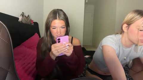 A video showing two young women with long hair, one in a burgundy sweater and the other in a grey crop top, sitting on a bed, focused on their smartphones.