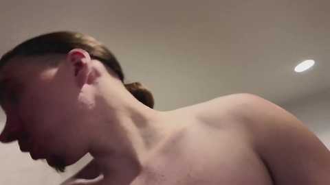 Video of a pale-skinned, young woman with short, light brown hair, slightly turned to the side, showing her bare shoulders and neck. The background is a simple, beige wall with a single, bright lightbulb overhead.