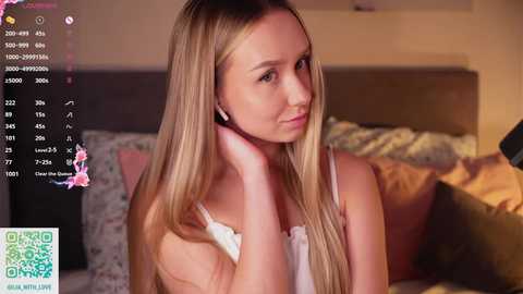 Video of a young Caucasian woman with long blonde hair, wearing a white tank top, smiling softly, in a cozy bedroom with floral decor.