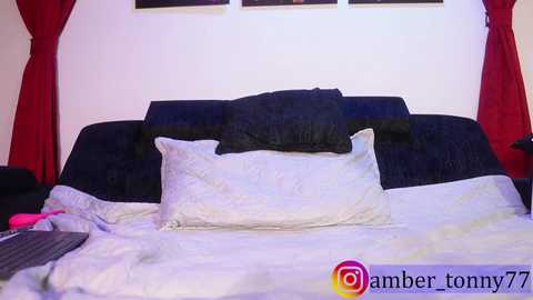 Video of a neatly made bed with white sheets and a black headboard. Red curtains frame the scene. A black pillow rests atop white pillows.