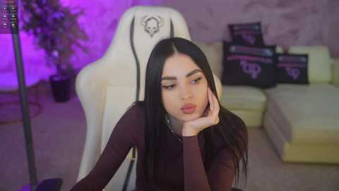 Video of a young woman with long black hair, wearing a burgundy top, leaning on her elbow in a white gaming chair. Background features a purple-lit room with a cream sofa and potted plants.