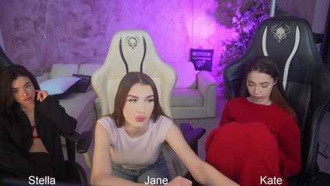 Video of three women in a gaming setup: Stella, Jane, and Kate. Stella wears a white top, Jane a red sweater, and Kate a black top. They sit in white gaming chairs with black skulls, surrounded by plants and purple lighting.