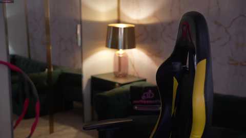A dimly lit, modern living room video featuring a black and yellow gaming chair, a lit table lamp, and a green couch, with a wooden floor and textured wallpaper in the background.