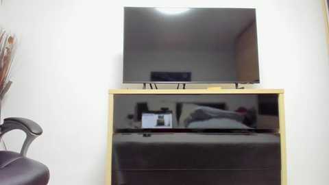 Video of a small, modern bedroom with a wall-mounted flat-screen TV, a wooden cabinet housing a black leather chair, and a neatly made bed with white sheets.