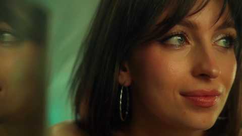 Video of a close-up of a woman with fair skin, green eyes, and straight dark hair, wearing hoop earrings, smiling gently, with a blurred background.