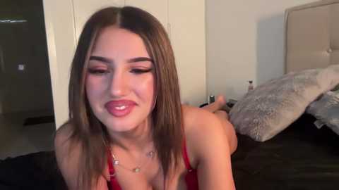Video of a young woman with long brown hair, light skin, and pink lipstick, lying on a bed, wearing a red bra. Background includes a beige headboard and a fluffy pillow.