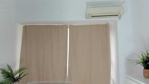 A video of a minimalist room featuring a beige curtain covering a window, an air conditioning unit on the wall above, and a potted plant on the right.