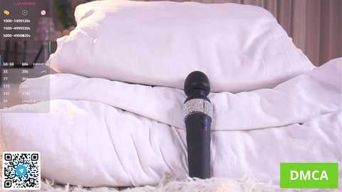 Video of a black vibrator with a diamond-encrusted handle lying on a white bed with rumpled sheets, surrounded by pillows, in a cozy bedroom setting.