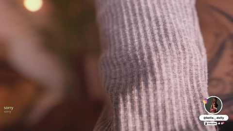 Media: A close-up video of a light gray ribbed sweater sleeve with a blurred background, featuring a small circular logo and text.