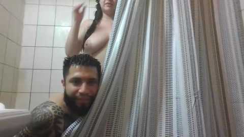 A video of a man with tattoos, wearing a black shirt, peeking through a shower curtain. Behind him, a woman with large breasts and long hair is visible.