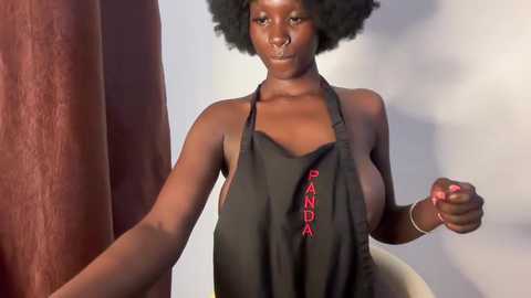 A CGI image of a dark-skinned woman with large breasts, wearing a black apron with \"PANDA\" in red letters, and a curly hairstyle. The background features a blurred, reddish-brown curtain.