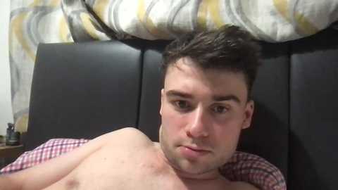 Video of a young, fair-skinned man with short, dark hair, lying shirtless on a bed with a black headboard. He has a neutral expression, and the background features a patterned, white and yellow pillow and a red and white checkered blanket.