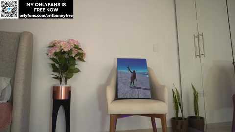 Media: Video of a minimalist bedroom featuring a white chair with a framed painting of a person on a beach, a plant, and a vase with pink flowers. Text overlay reads, \"MY ONLYLYNS IS FREE NOW.\