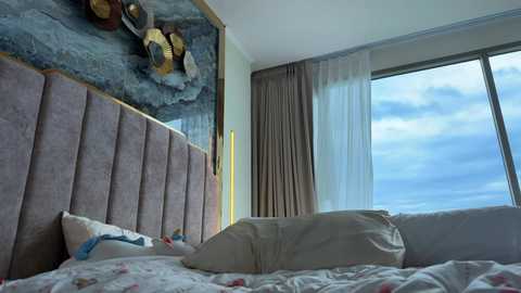 Video of a modern bedroom with a large, light gray tufted headboard, beige pillows, and floral bedspread. Large window with sheer curtains, allowing a view of a cloudy sky. Decorative wall art above the headboard.