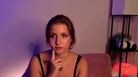 Video of a young Caucasian woman with light skin and brown hair, wearing a black top, seated indoors with a thoughtful expression. She touches her chin with her finger. Background includes a beige sofa, a potted plant, and a black lamp.