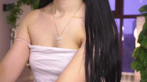 Video of a fair-skinned woman with long black hair, wearing a strapless white top, and a silver cross necklace. Background features green plants and a window with purple lighting.