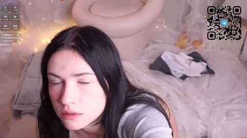Video of a young woman with long black hair, fair skin, and closed eyes, lying on a white sheet in a messy room with a toilet and scattered clothes.