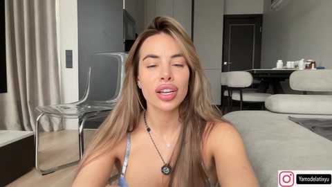 Video of a light-skinned woman with long, straight brown hair, wearing a blue bra, and a necklace, sitting in a modern living room with a clear chair, beige sofa, and kitchen in the background.