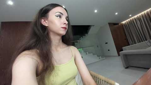 Video of a fair-skinned, slender woman with long, wavy brown hair in a light yellow spaghetti-strap top, standing in a modern, minimalist living room with white walls, wooden furniture, and a staircase.