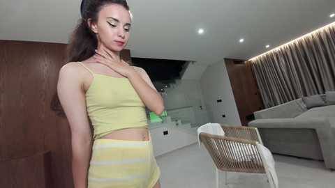 Video of a slender woman with long dark hair in a yellow crop top and striped skirt, standing in a modern living room with a grey couch and wooden walls.