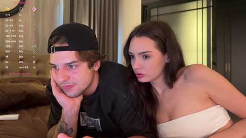 Video of a young man with tattoos in a black hat and shirt, leaning on a woman with long hair and a white strapless dress, in a modern bedroom.