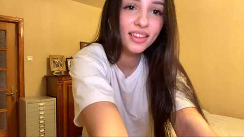 A video of a young woman with long, dark hair, wearing a white t-shirt, smiling in a cozy, yellow-walled bedroom with a wooden door, desk, and framed pictures.