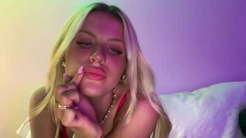 Video of a blonde woman with long hair, wearing a red top, making a peace sign with her finger, against a purple-lit background.