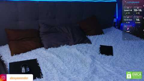 Video of a dimly lit bedroom with a plush, white faux fur blanket, a brown pillow, and a black laptop on a bed, under a blue LED strip light.