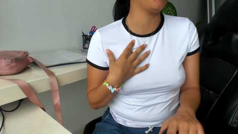 Video of a young woman with medium skin tone, wearing a white T-shirt, seated at a desk. She has black hair and a colorful bracelet on her wrist. Her left hand is placed on her chest. Background shows a pink bag, a laptop, and office supplies.