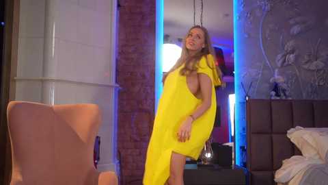 Video of a slender, fair-skinned woman with long blonde hair wearing a bright yellow dress, standing in a modern, dimly lit room with exposed brick walls, a plush armchair, and a bed.
